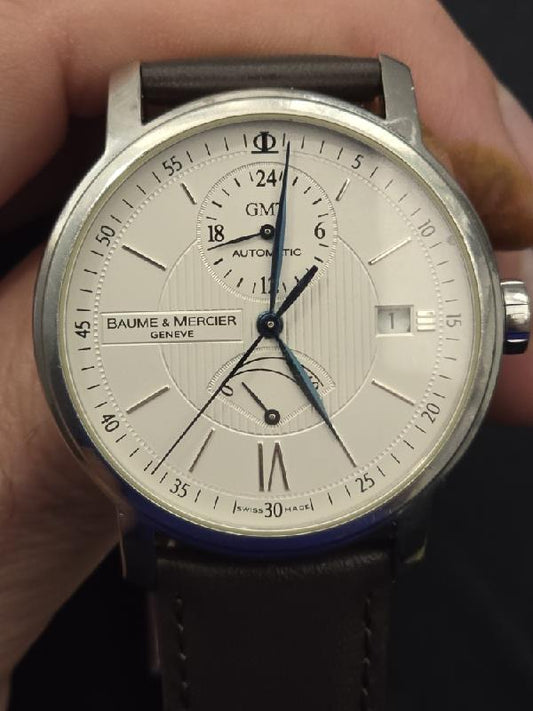 Baume & Mercier GMT automatic dual time power reserve watch with black dial, silver case, and leather strap.
