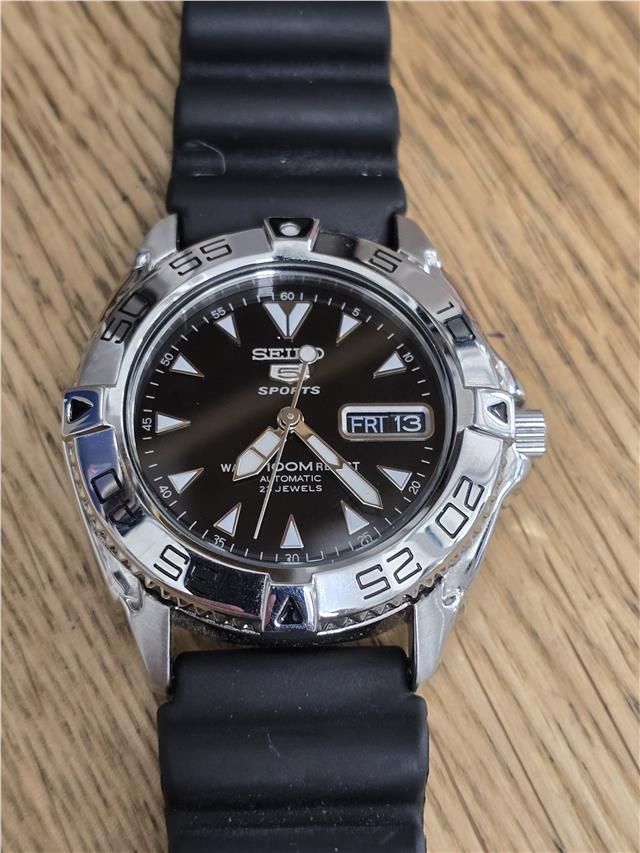 Seiko 5 Sports automatic watch with boxed papers, featuring a stainless steel strap and black dial, displayed on a white background.